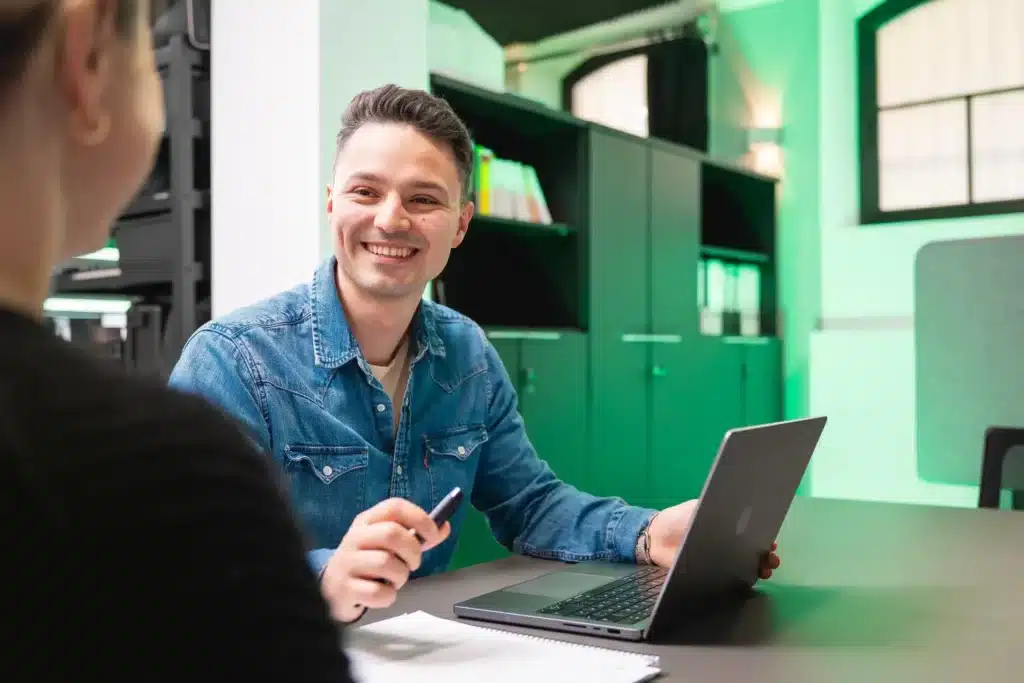 Eine Person im Jeanshemd sitzt mit Laptop und Notizblock an einem Tisch und lächelt, während sie mit jemandem in der Nähe SEO-Strategien bespricht. Das grünliche Licht wirft einen warmen Schein auf die Regale im Hintergrund.