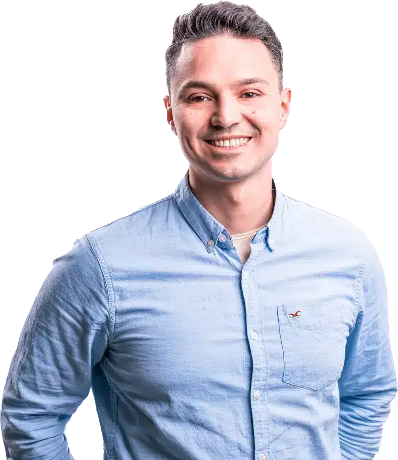 Ein lächelnder Mann in einem hellblauen Hemd mit hochgekrempelten Ärmeln und einem kleinen Logo auf der linken Seite steht selbstbewusst mit einer Hand auf der Hüfte. Mit kurzen, dunklen Haaren blickt der SEO-Experte vor einem schlichten Hintergrund in die Kamera.