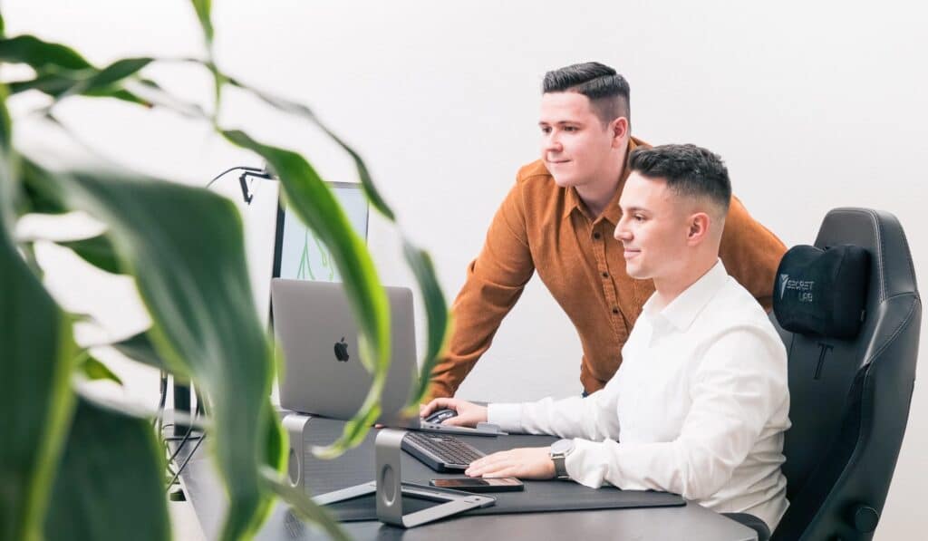 In einem eleganten, modernen Büro sitzt ein SEO-Agentur-Mitarbeiter fleißig am Laptop, während ein Kollege daneben steht. Beide schauen konzentriert auf den Bildschirm. Eine üppige Pflanze im Vordergrund bringt einen Hauch von Grün in die produktive Umgebung.