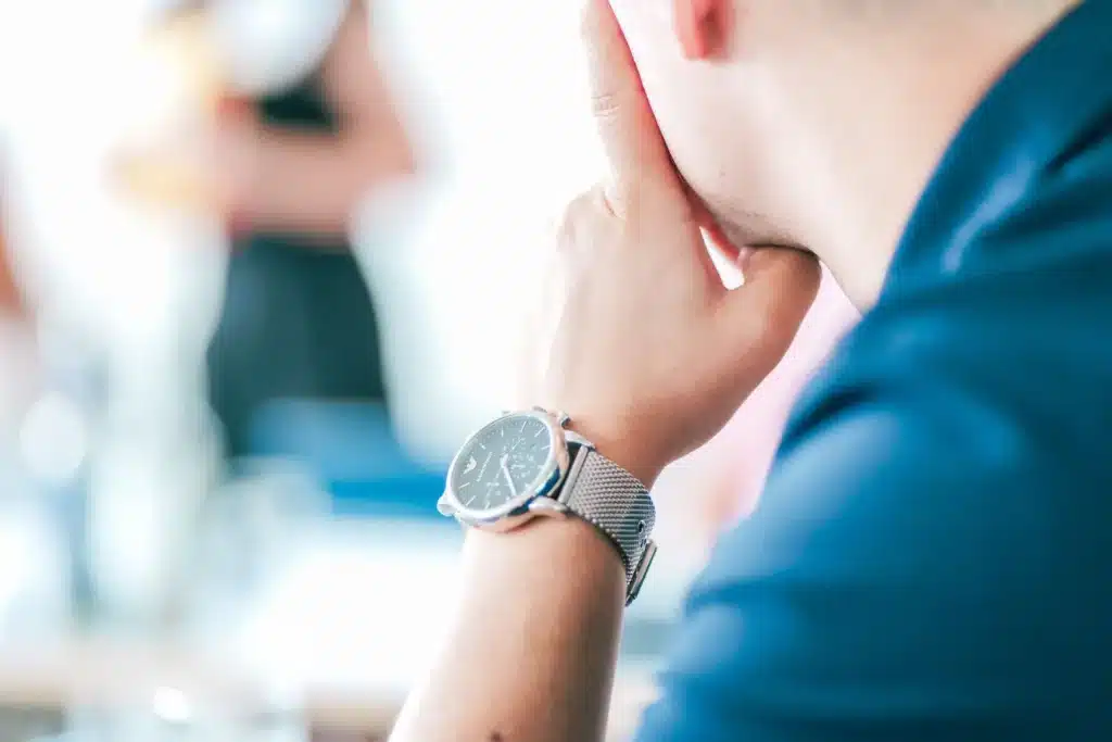 Eine Person in blauem Hemd, die möglicherweise über SEO-Strategien in Nürnberg nachdenkt, stützt ihr Kinn auf die Hand. Eine silberne Uhr glänzt an ihrem Handgelenk, während der Hintergrund verschwimmt und undeutliche Figuren in der Ferne verschwinden.