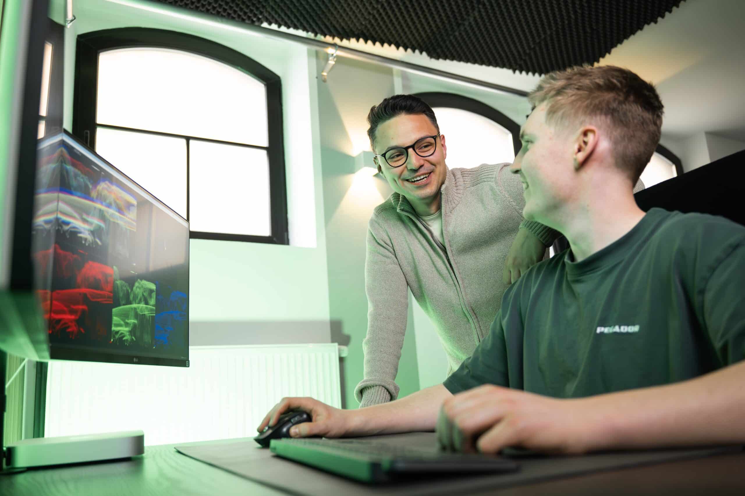 In einer modernen Büroumgebung arbeiten zwei Personen zusammen: Eine sitzt an einem Schreibtisch und konzentriert sich auf SEO-Strategien mit farbenfrohen Bildern auf dem Bildschirm, während die andere lächelnd daneben steht. Sanfte Beleuchtung und große Fenster fördern die kollaborative Atmosphäre.