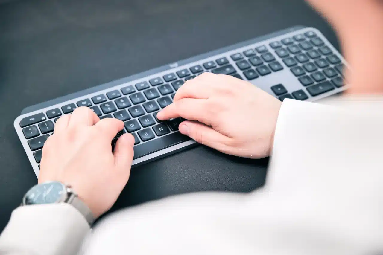 Nahaufnahme einer Person, die mit beiden Händen auf einer schwarzen Tastatur tippt und möglicherweise eine SEO-Strategie entwickelt. Die Person trägt ein weißes Hemd und eine Uhr am linken Handgelenk vor einem dunklen Hintergrund.